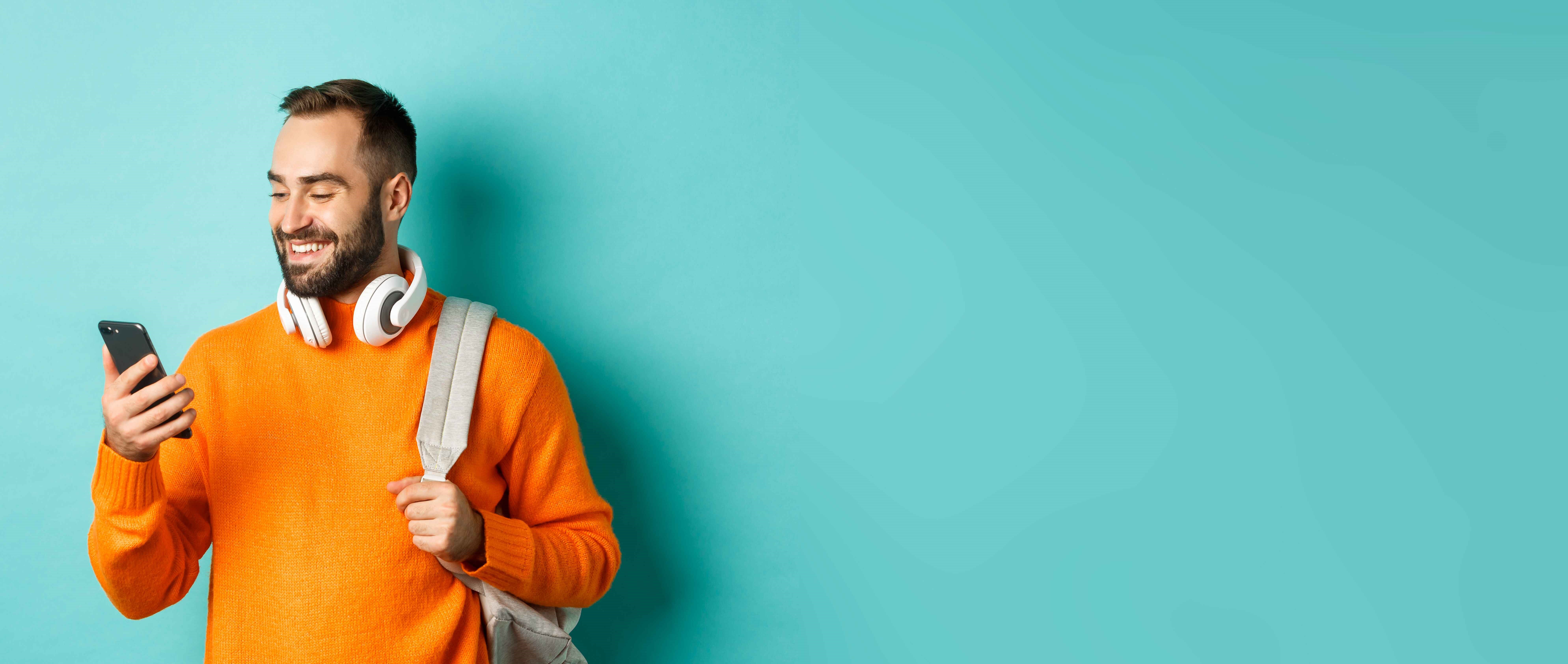 caucasian-man-with-headphones-backpack-looking-phone-reading-message-smiling-standing-ove (1)